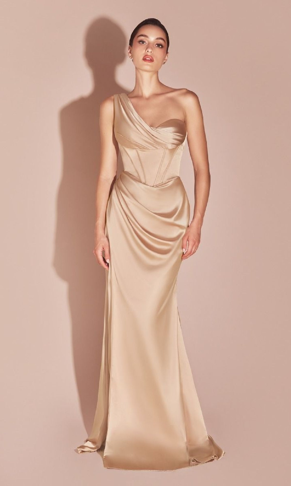A woman wears the Ladivine 7448 One-Shoulder Lace-Up Prom Dress, a champagne satin floor-length gown with a corset bodice, standing against a plain beige background.